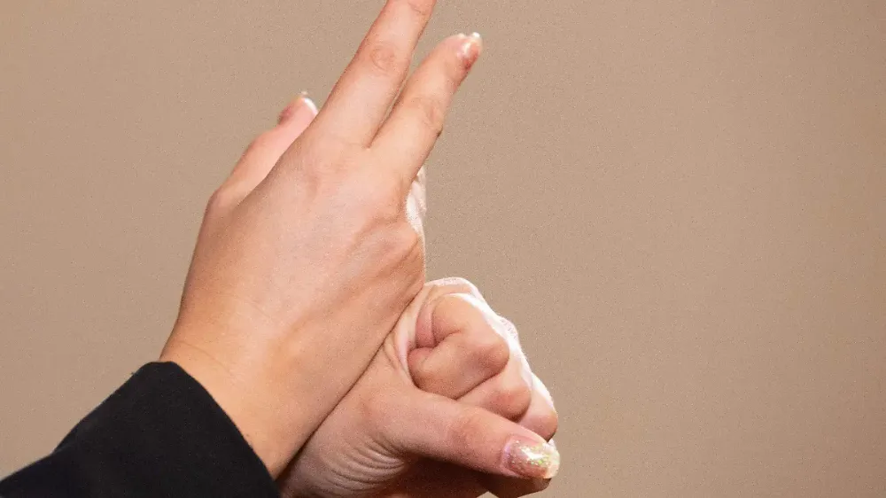Primer plano de dos manos haciendo una seña en lengua de señas sobre un fondo beige, con dedos extendidos hacia arriba